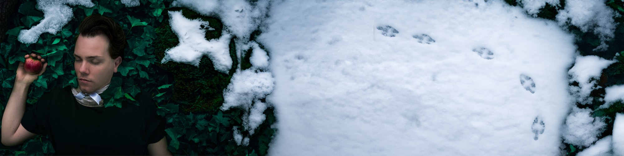 Mika Maria Mausshammer liegt im Efeu und hält einen roten Apfel, daneben frischer Schnee mit Vogelspuren